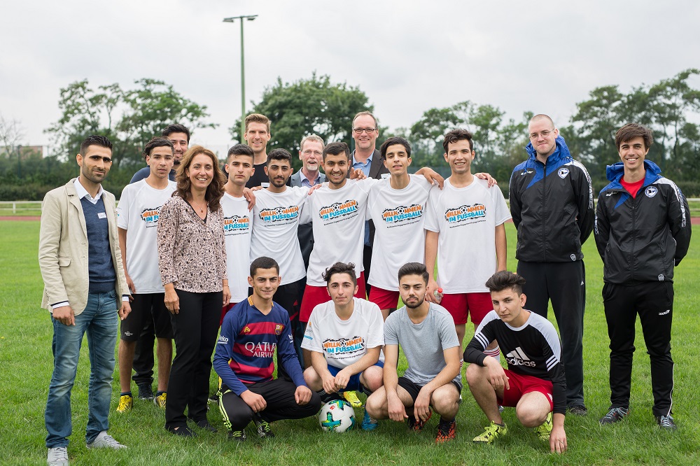 Eine Gruppe junger Männer und Frauen, hauptsächlich im Teenageralter, steht auf einem Fußballfeld und lächelt in die Kamera. Sie tragen weiße T-Shirts mit der Aufschrift "Willkommen im Fußball" und Sportkleidung. Einige Erwachsene stehen ebenfalls in der Gruppe und wirken wie Betreuer oder Trainer. Im Vordergrund knien einige Jugendliche um einen Fußball.