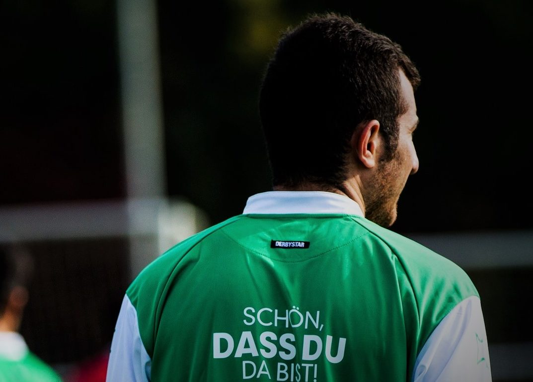 Ein Fußballspieler steht mit dem Rücken zum Betrachter da. Auf seinem grünen Trikot steht in großen weißen Buchstaben "SCHÖN, DASS DU DA BIST!".