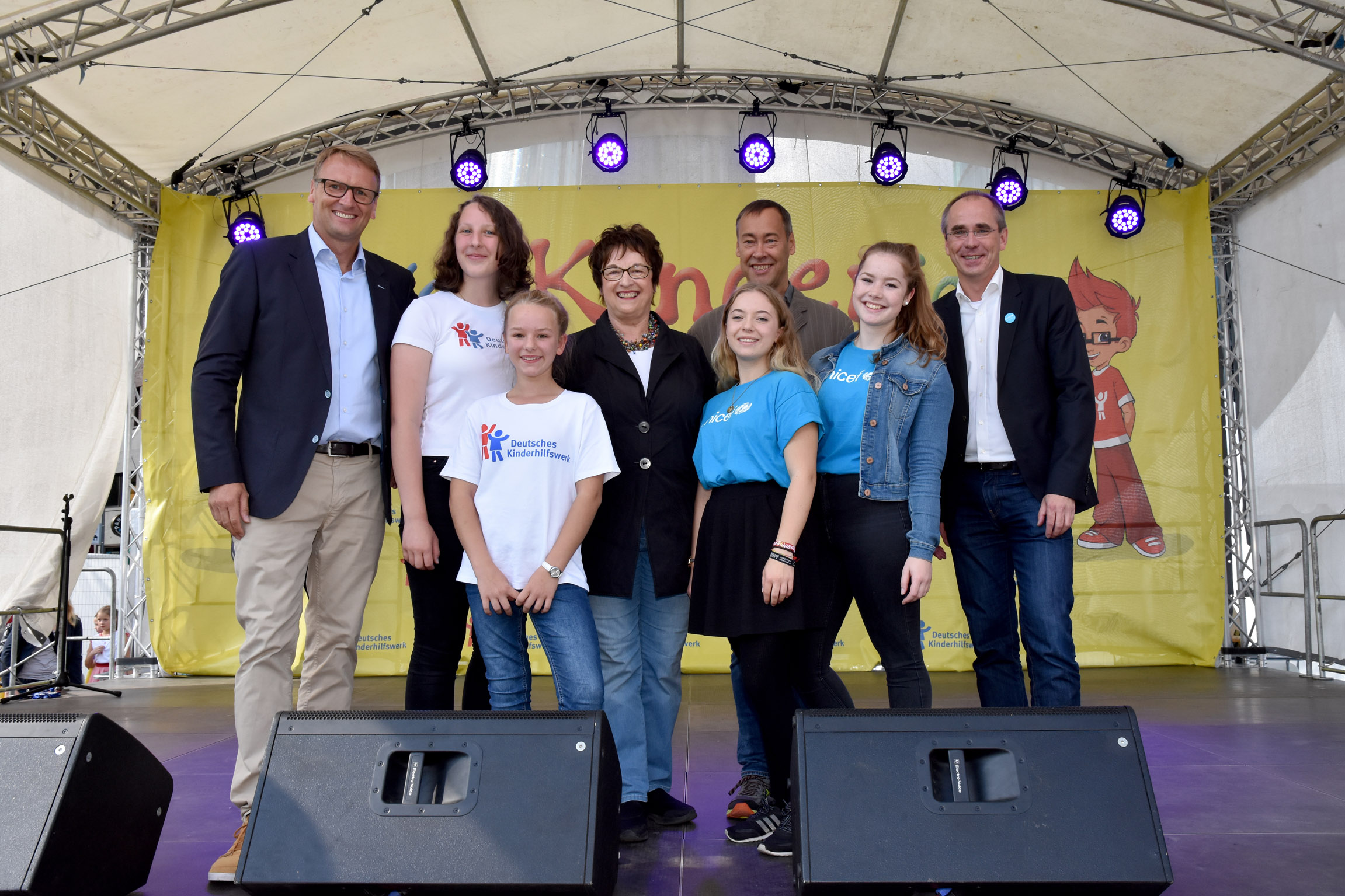 Eine Gruppe von sieben Personen steht auf einer Bühne vor einer Wand mit dem Aufdruck "Deutsches Kinderhilfswerk". Die Personen sind unterschiedlich gekleidet, darunter ein Mann im Anzug, Frauen in Jeans und T-Shirts sowie zwei junge Mädchen. Im Hintergrund ist ein Banner mit einem kindlichen Motiv zu sehen.
