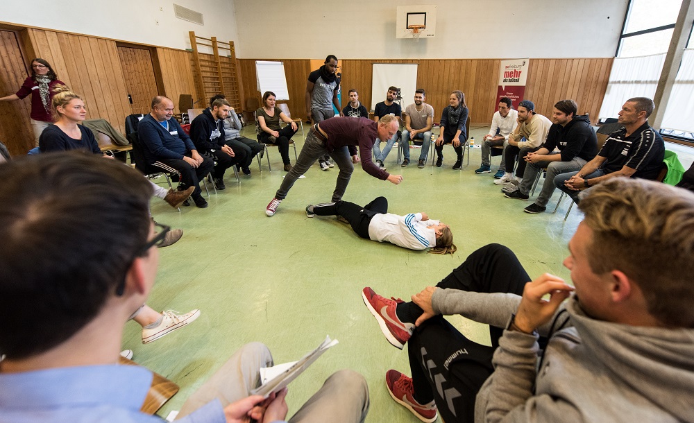 Eine Gruppe von Menschen sitzt im Kreis in einer Turnhalle. In der Mitte liegt eine Person auf dem Rücken, während ein anderer Teilnehmer in ihre Richtung geht, möglicherweise um zu helfen oder eine Übung zu erklären. Im Hintergrund sind weitere Teilnehmer sichtbar, die die Szene beobachten. Das Bild illustriert einen Workshop der DFL Stiftung zum Thema Konfliktbewältigung im Fußball.