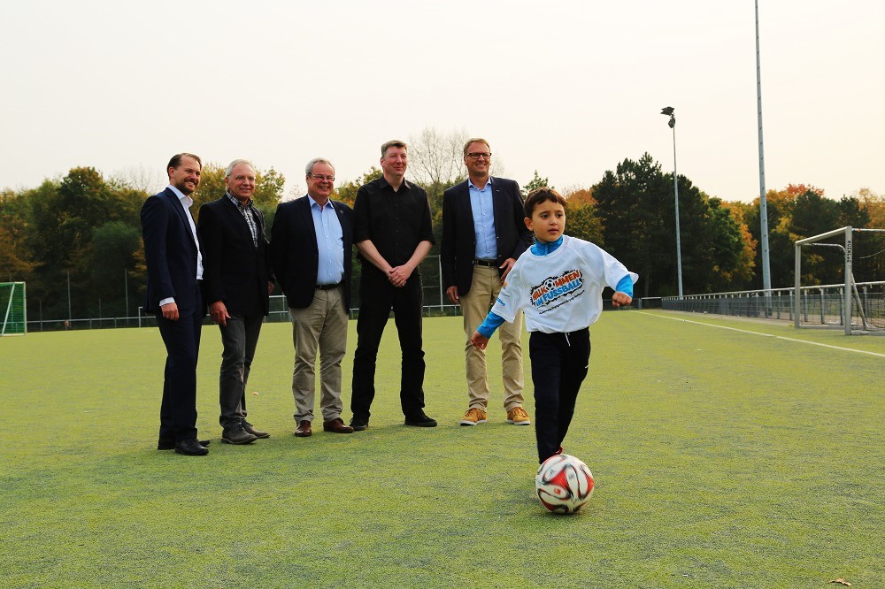 Junge Fußballspieler dribbelt auf einem grünen Kunstrasenplatz, umgeben von vier Männern in Anzügen, im Rahmen des Willkommensbündnisses "Willkommen in Fußball".