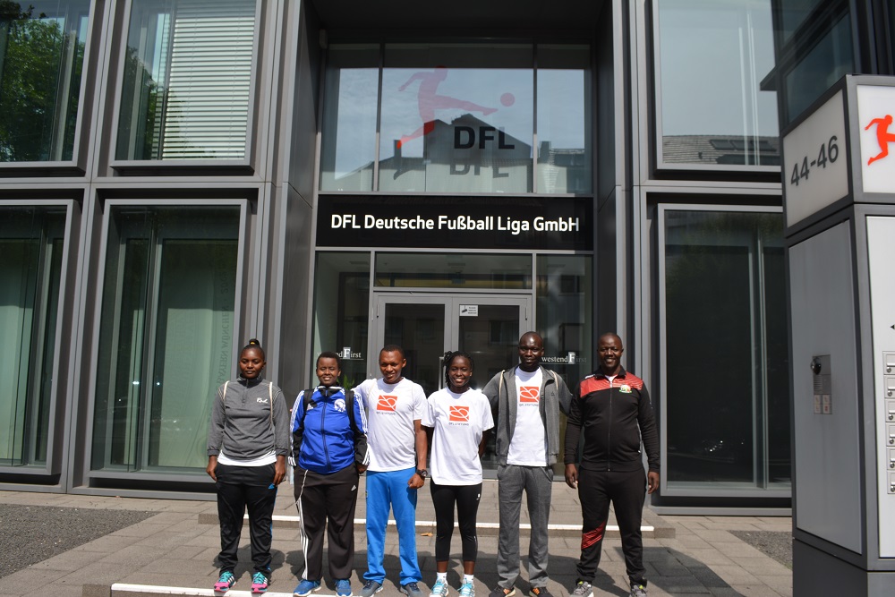 Eine Gruppe von sieben jungen Menschen steht vor dem modernen Glasgebäude der DFL Deutsche Fußball Liga GmbH. Sie tragen Trainingskleidung und schauen in die Kamera.