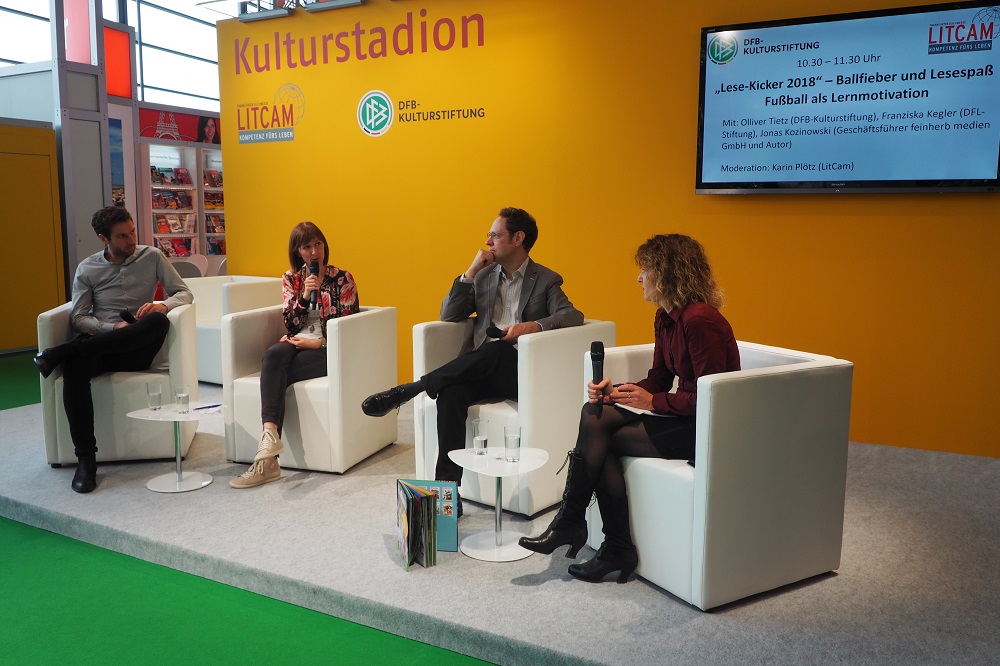 Eine Gesprächsrunde mit vier Personen auf weißen Sesseln vor einem großen Bildschirm mit dem Aufdruck "Lese-Kicker 2018 – Ballfieber und Lesespaß – Fußball als Lernmotivation". Im Hintergrund ist ein grün gestrichenes Mauerwerk zu sehen.