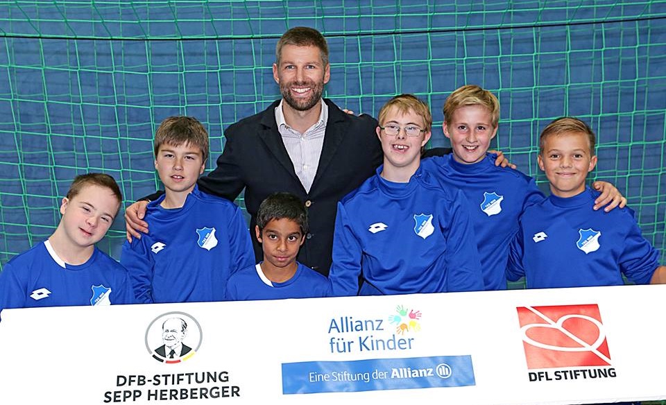 Thomas Hitzlsperger umarmt lächelnd eine Gruppe von jungen Fußballspielern in blauen Trikots. Im Hintergrund ist ein Banner mit den Logos der DFL Stiftung, der Allianz Stiftung und der DFB Stiftung Sepp Herberger zu sehen.
