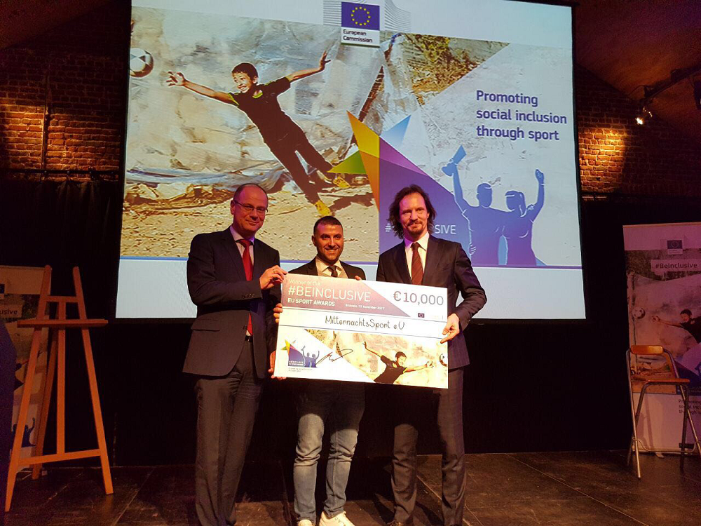 Tibor Navracsics, EU-Kommissar für Bildung, Kultur, Jugend und Sport, und Estlands Kulturminister Indrek Saar überreichen Ismail Öner, Initiator von MitternachtsSport, einen Scheck über 10.000 Euro im Rahmen des DFL Stiftung MitternachtsSport Awards. Im Hintergrund ist eine Projektion mit dem Slogan "Promoting social inclusion through sport" zu sehen.