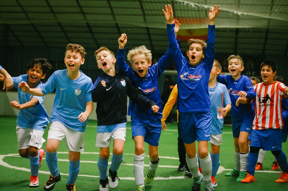 Jubelnde Kinderfußballmannschaft in blauen und orangen Trikots auf einem grünen Kunstrasenfeld.
