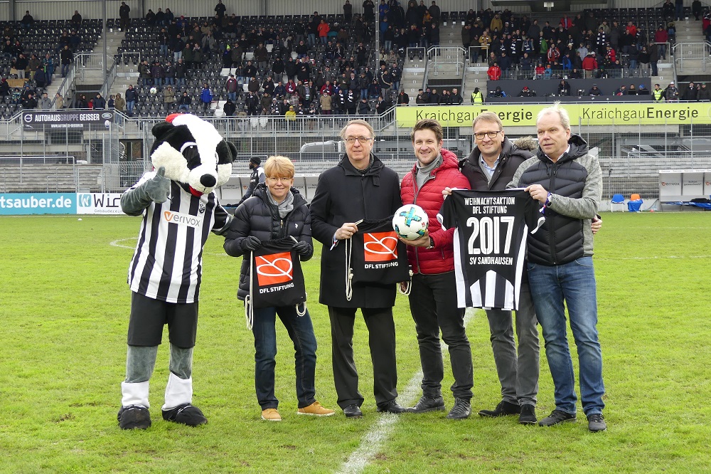 Gruppe von Menschen steht auf einem Fußballfeld, darunter der DFL Stiftung-Maskottchen, mit einer Banner mit der Aufschrift "Weihnachtsaktion DFL Stiftung 2017 SV Sandhausen".