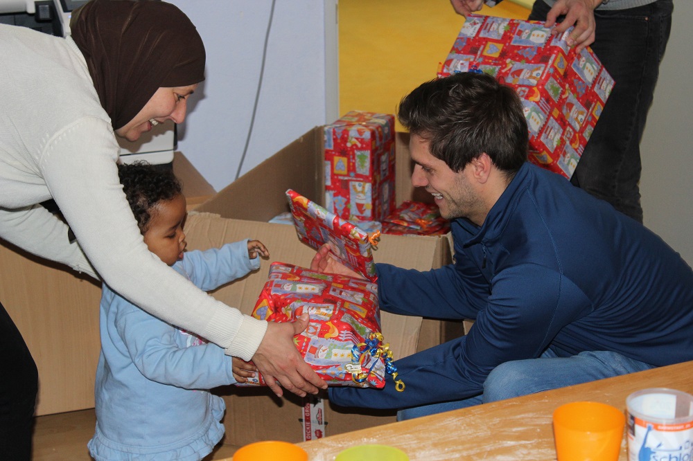 Ein Kind und ein Mann reichen sich freudig Winterpakete über einen Tisch. Im Hintergrund eine Frau, die ebenfalls Pakete verteilt. Die Pakete sind bunt in rotes Geschenkpapier verpackt.