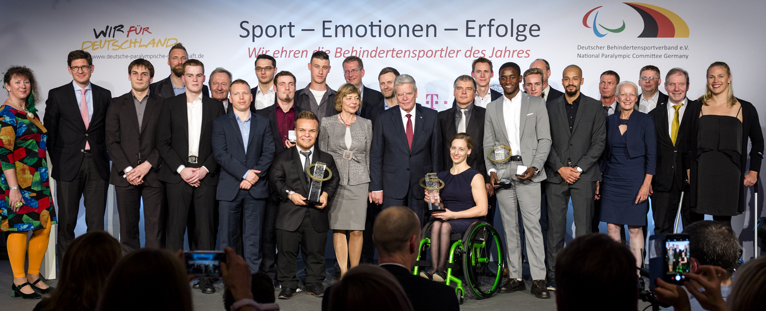 Eine Gruppe von Sportlern und Funktionären steht auf einer Bühne. Im Vordergrund halten zwei Sportler im Rollstuhl Pokale hoch. Im Hintergrund sind weitere Sportler und Ehrengäste zu sehen. Auf einem Banner im Hintergrund steht "Ehrung der Behindertensportler des Jahres".
