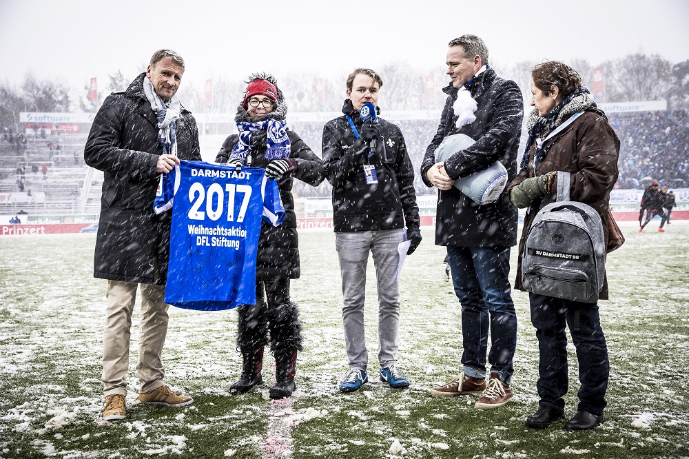 Eine Gruppe von Personen steht bei Schneefall auf dem Rasen eines Fußballstadions. In den Händen halten sie eine blaue Fahne mit der Aufschrift "2017 - Weihnachtsaktion - DFL Stiftung". Im Hintergrund ist das Stadion und die Zuschauertribüne erkennbar.