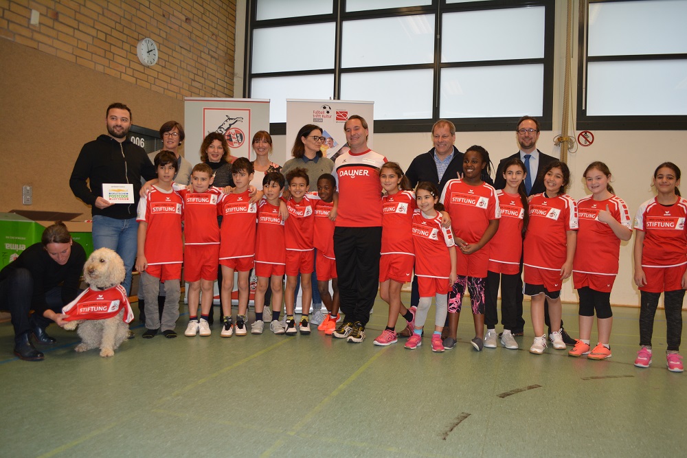 Eine Gruppe von Kindern in roten T-Shirts mit dem Aufdruck "STIFTUNG" steht in einer Sporthalle. Sie nehmen an dem Projekt "Fußball trifft Kultur" der DFL Stiftung teil. Im Hintergrund stehen Betreuer und Organisatoren. Ein Teddybär mit dem Schriftzug "STIFTUNG" sitzt vor der Gruppe.