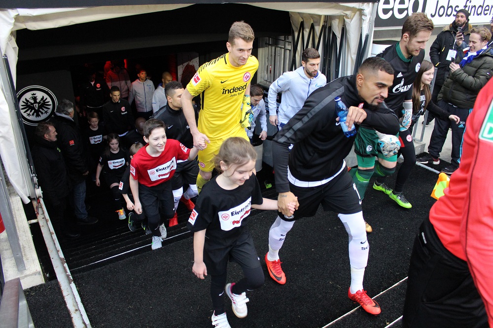 Ein Fußballspieler in gelbem Trikot führt Kinder in roten Trikots aus einem Tunnel auf den Rasen. Im Hintergrund stehen weitere Kinder und Betreuer. Das Bild zeigt eine Kooperation zwischen der DFL Stiftung und der Eintracht Frankfurt Pausenliga.