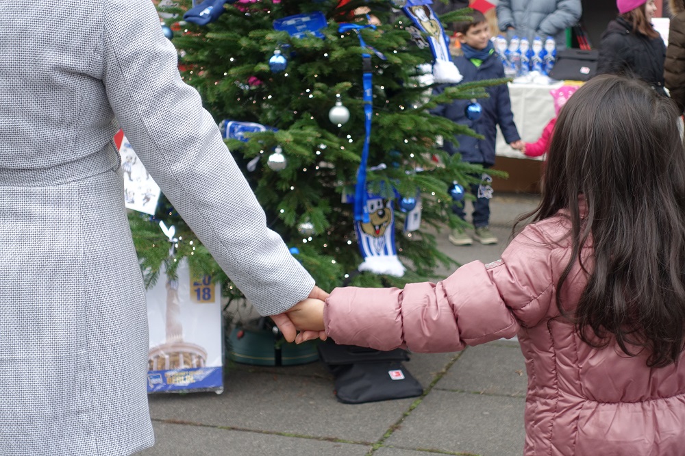 Zwei Kinder halten sich an den Händen und stehen vor einem geschmückten Weihnachtsbaum, im Hintergrund stehen weitere Personen und Tische.
