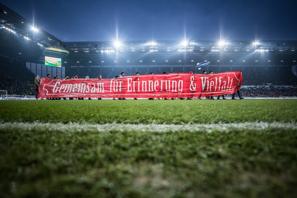 Ein Banner mit der Aufschrift "Gemeinsam für Erinnerung & Vielfalt" wird vor einer Fußballstadionkulisse gezeigt. Im Hintergrund sind die Lichter und Tribünen der OPEL ARENA in Mainz zu sehen. Das Bild wurde anlässlich des Erinnerungstages im Januar 2017 aufgenommen.