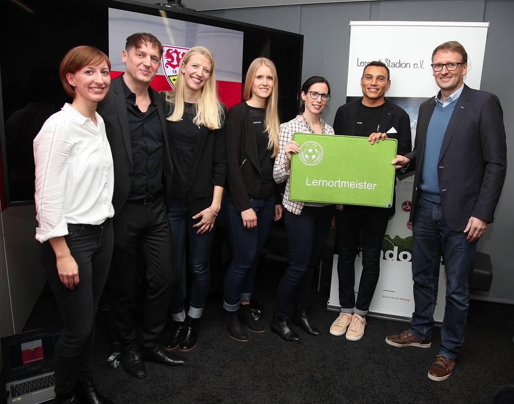 Eine Gruppe von sieben Personen steht lächelnd zusammen und hält eine grüne Plakette mit der Aufschrift „Lernmeister“ hoch. Im Hintergrund ist ein Banner mit dem Logo des VfB Stuttgart zu sehen.