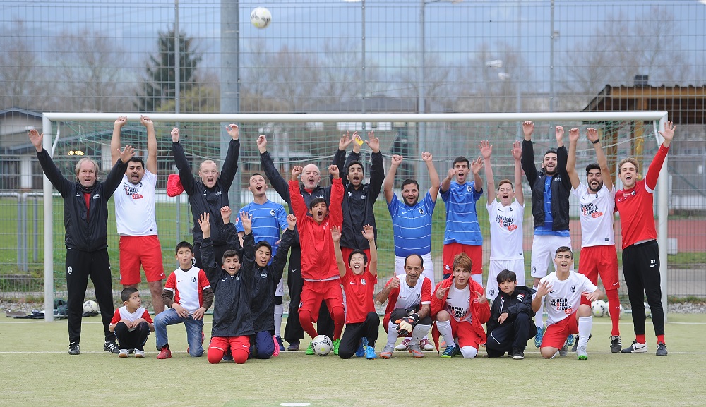 Eine Gruppe von Menschen, darunter Fußballspieler und Helfer, steht auf einem Fußballfeld vor einem Tor und reckt die Arme in die Höhe. Einige tragen Fußballtrikots, andere Sportkleidung oder Jacken. Das Bild zeigt eine Aktion der DFL Stiftung, bei der Sportbekleidung für Flüchtlinge gespendet und ein Fußballspiel veranstaltet wurde.