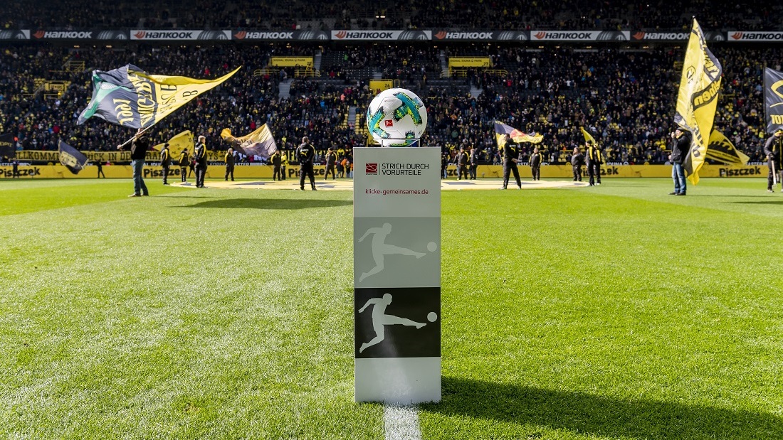 Fußballstadion, im Vordergrund eine Werbetafel mit dem Aufdruck "Strich durch Vorurteile" und einem stilisierten Fußballspieler, im Hintergrund Fans im Stadion.