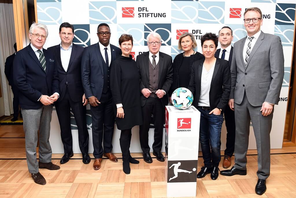 Eine Gruppe von acht Personen steht lächelnd vor einem Podest mit einem Fußball darauf. Im Hintergrund ist ein Banner mit dem Logo der DFL Stiftung und der Bundesliga zu sehen.