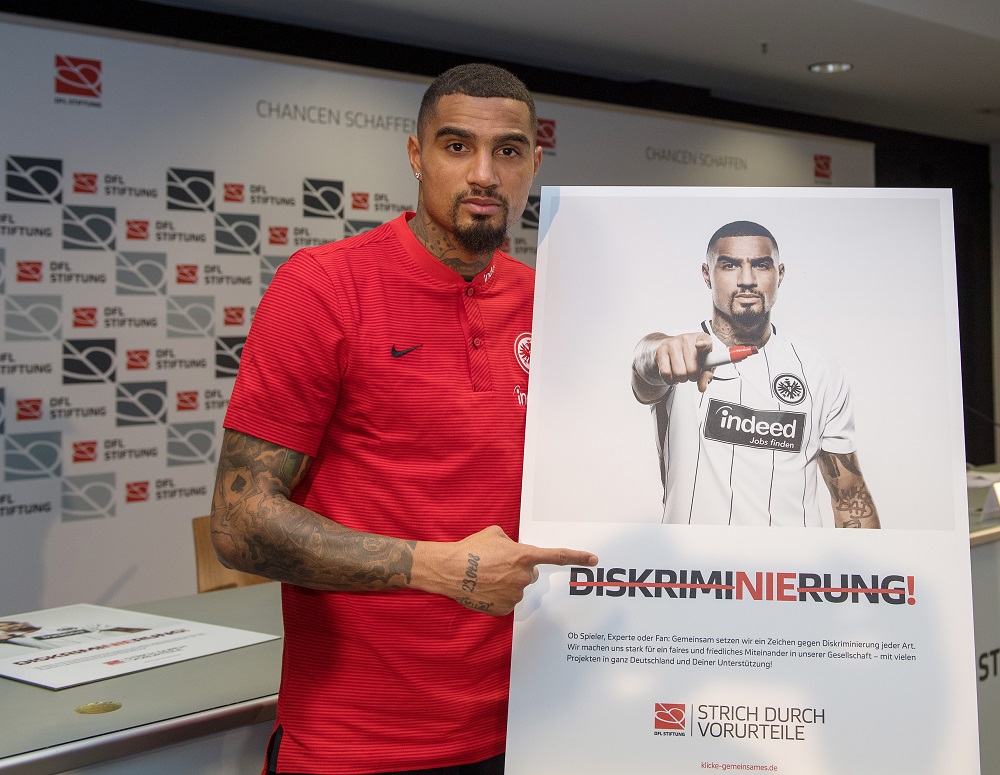 Ein Mann mit Tattoos und rotem Polohemd hält ein großes Plakat mit dem Aufdruck "Diskriminierung!" und dem Slogan "Strich durch Vorurteile" in der Hand. Das Plakat zeigt ein schwarz-weißes Foto einer Person mit erhobener Hand, die ein Zeichen gegen Diskriminierung macht. Im Hintergrund ist ein Banner mit dem Logo der DFL Stiftung zu sehen.