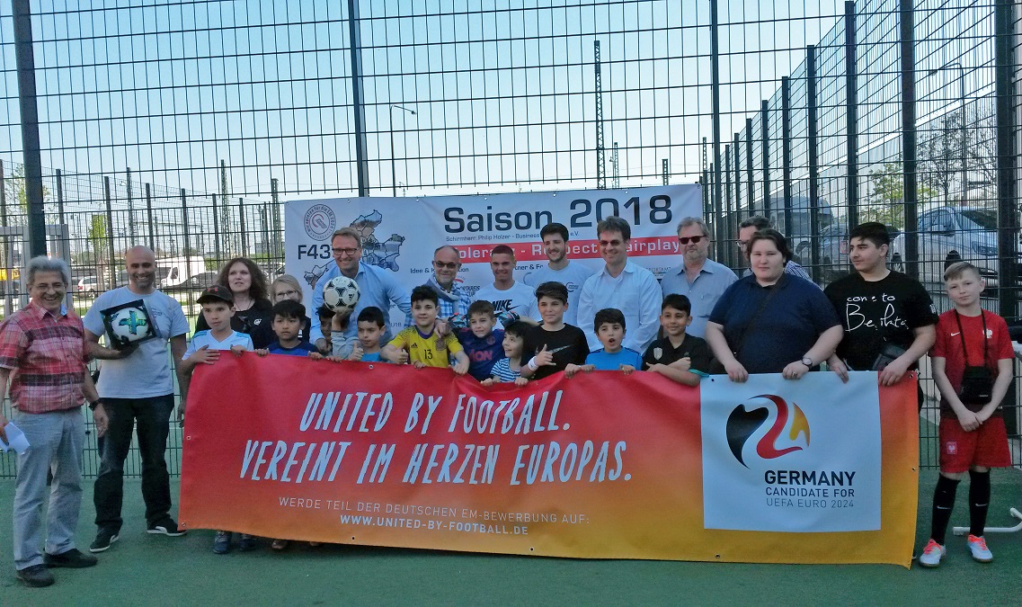 Eine Gruppe von Kindern und Jugendlichen steht auf einem Bolzplatz vor einem Banner, das "UNITED BY FOOTBALL. VEREINT IM HERZEN EUROPAS." zeigt. Im Hintergrund ist die Aufschrift "Saison 2018" zu sehen. Das Banner wirbt für die Bewerbung Deutschlands als Gastgeber der UEFA EURO 2024.
