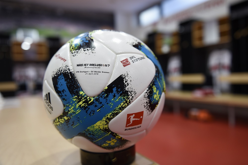 Fußballball mit dem Aufdruck "WAS IST INKLUSION?" und dem Logo der DFL Stiftung. Im Hintergrund ist ein Raum mit Sitzplätzen und Regalen zu erkennen.