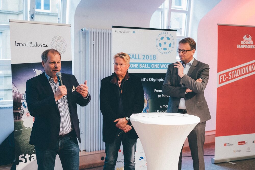Drei Männer in Anzügen stehen auf einer Veranstaltung im Freien vor einem Banner mit dem Schriftzug "BALL 2018 - ONE BALL, ONE WORLD". Der Mann in der Mitte hält ein Mikrofon und spricht. Im Hintergrund ist das Logo "Spirit of Football" zu sehen.