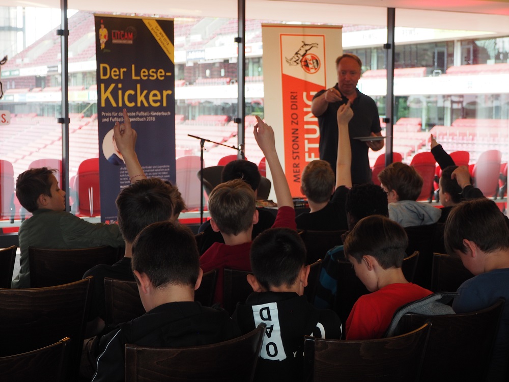 Ulf Blanck steht vor einer Gruppe Kinder und liest aus dem Buch "Die drei ??? - Der Fußball Roboter" vor. Im Hintergrund ist ein Stadion zu sehen.