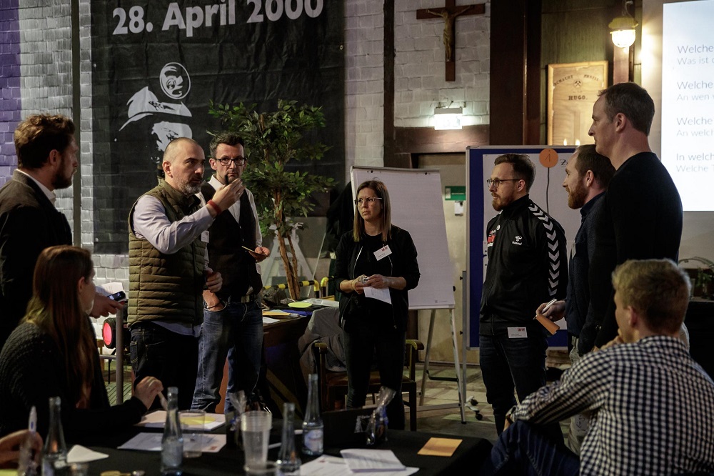 Eine Gruppe von Personen sitzt und steht in einem hellen Raum zusammen und diskutiert lebhaft. Einige halten Stifte und Notizblöcke, während sie sich auf einen Tisch mit Getränken und Papieren konzentrieren. Im Hintergrund ist eine Tafel oder eine Präsentationsfläche zu sehen. Die Atmosphäre ist konzentriert und gemeinschaftlich.