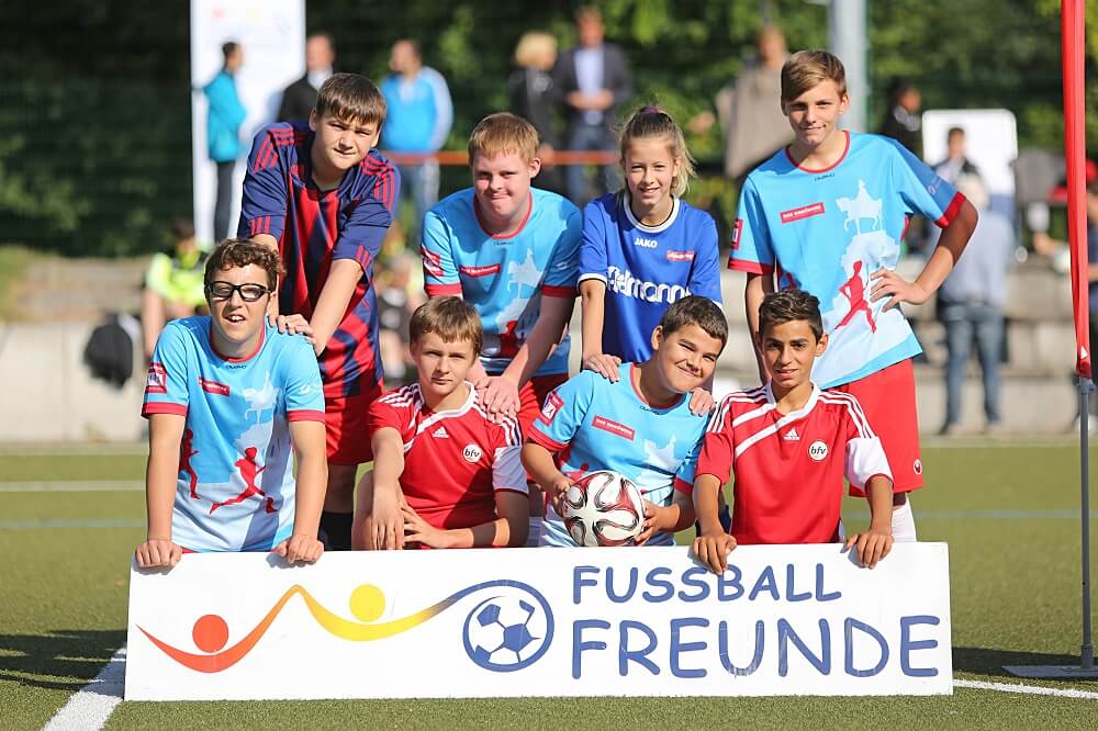 Eine Gruppe von Kindern und Jugendlichen in Fußballtrikots steht auf einem grünen Rasen vor einem Banner mit der Aufschrift "FUSSBALLFREUNDE". Sie lächeln in die Kamera und halten einen Fußball. Im Hintergrund sind weitere Personen und Sportgeräte erkennbar.