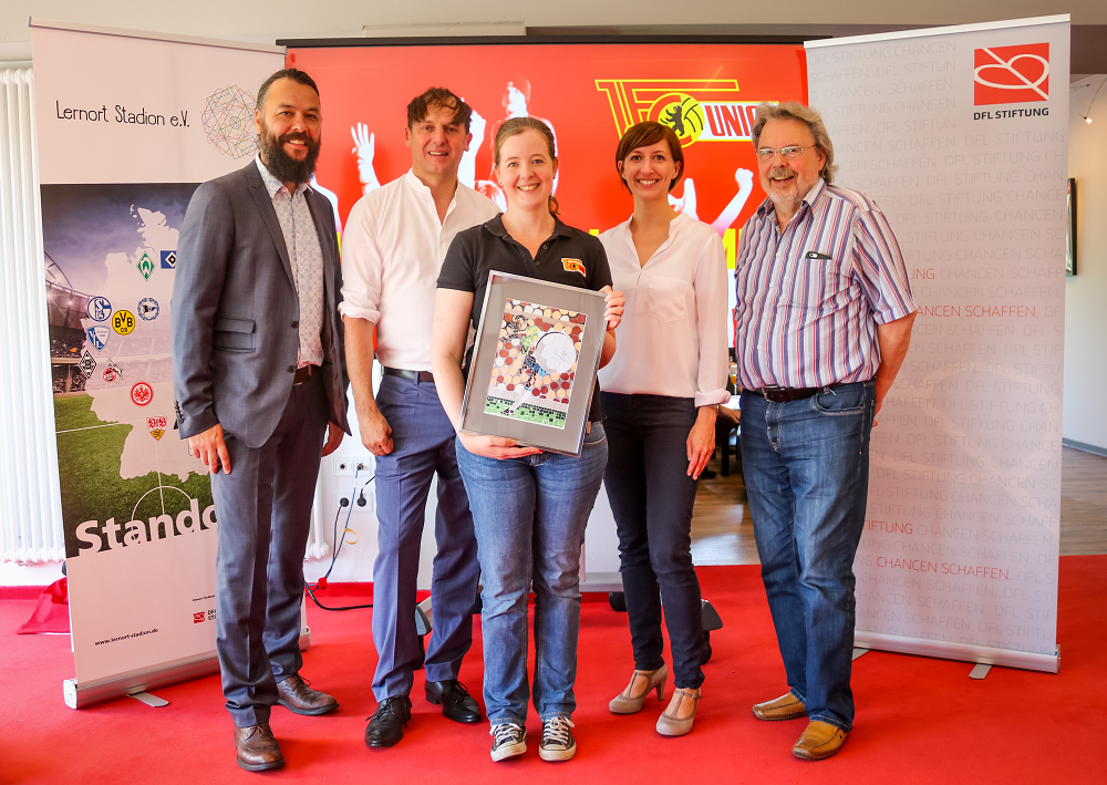 Eine Gruppe von vier Personen steht lächelnd vor einem Banner mit dem Logo der DFL Stiftung und dem Logo des 1. FC Union Berlin. Im Zentrum hält eine Frau eine quadratische Plakette mit einem Fußballmotiv in den Händen. Im Hintergrund ist ein Banner mit verschiedenen Fußballvereinslogos zu sehen, was auf eine Partnerschaft im Bereich Sport und soziales Engagement hindeutet.