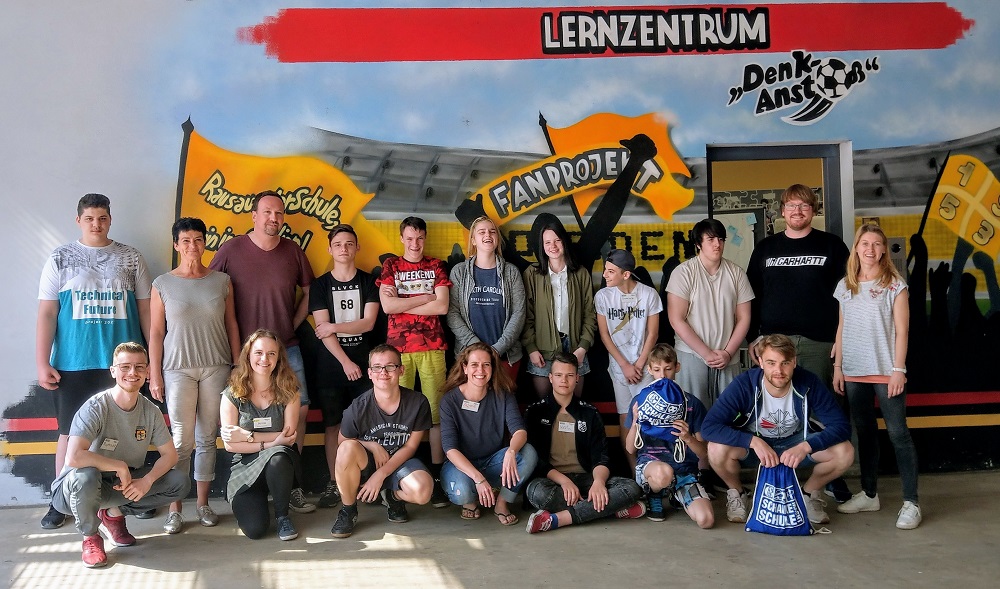 Eine Gruppe Jugendlicher steht und hockt vor einer Wand mit Graffiti-Motiven, die verschiedene Lernzentren und das Logo des Projekts "Lernort Stadion" zeigt. Im Hintergrund ist ein Stadion zu sehen. Die Jugendlichen sind Teil des Projekts "All Inclusive", das von den Lernzentren "Schalke macht Schule" und "Denk-Anstoß" durchgeführt wird.