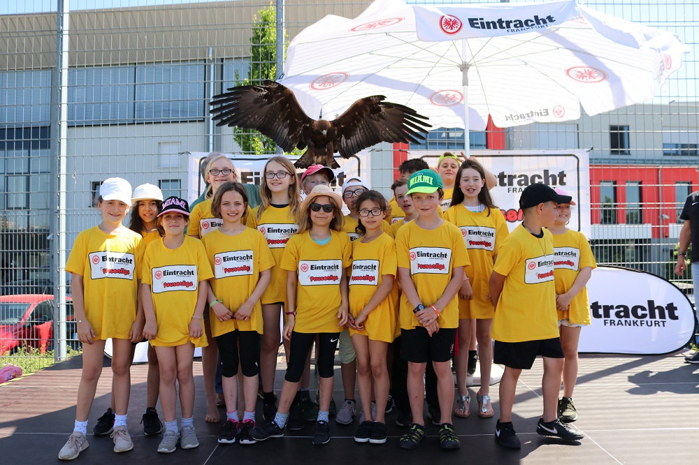Eine Gruppe von Kindern und Jugendlichen in gelben T-Shirts mit dem Eintracht Frankfurt Logo steht vor einer großen, weißen Pappfigur eines Raubvogels. Im Hintergrund ist ein Gebäude und ein Werbebanner für "tracth Frankfurt" zu sehen.