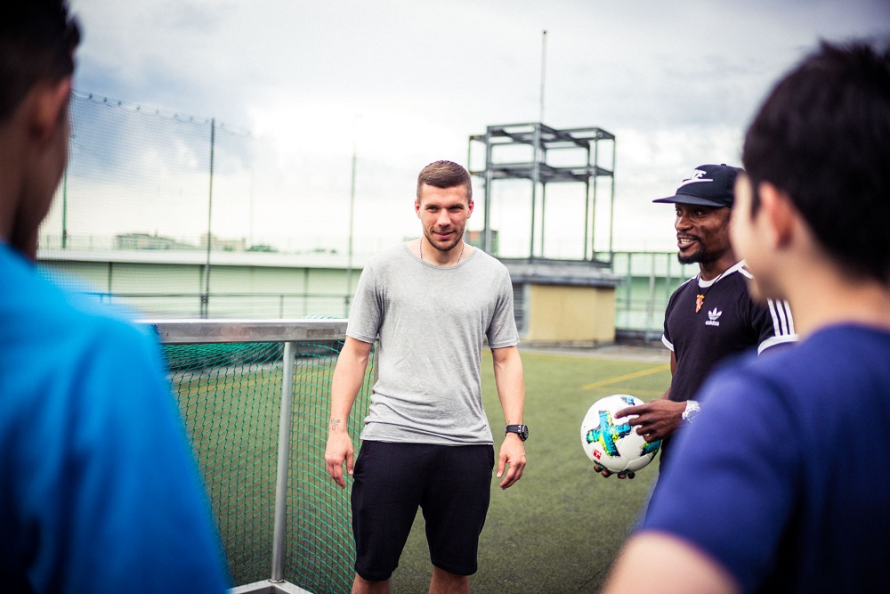 Fußballspieler Lukas Podolski umgeben von Jugendlichen auf einem Fußballplatz, im Gespräch und mit einem Fußball in der Hand.
