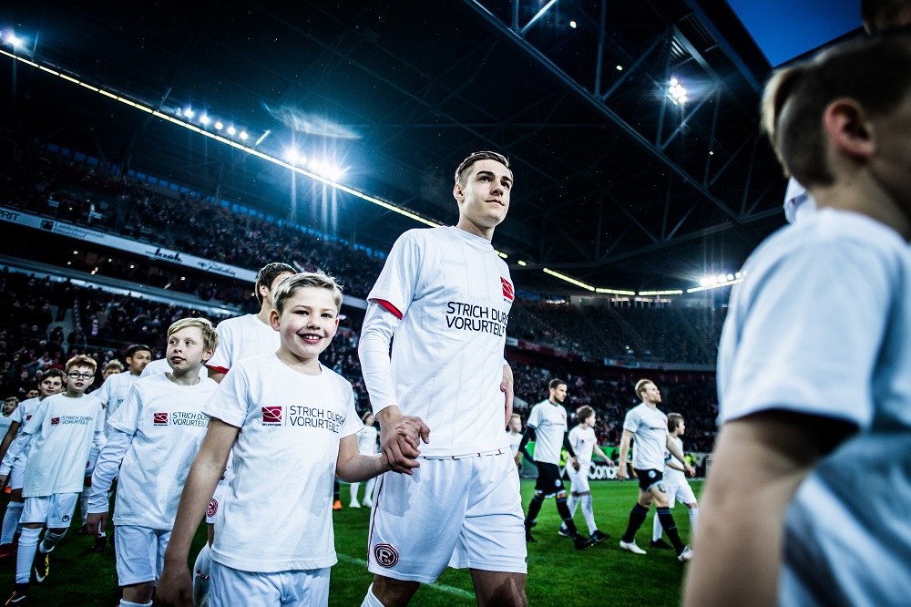 Fußballspieler Florian Neuhaus von Fortuna Düsseldorf geht Hand in Hand mit einem Kind aus der Begleitung auf das Spielfeld, im Hintergrund weitere Spieler und Fans im Stadion. Auf den Trikots ist der Aufdruck "Strich durch Vorurteile" zu sehen.