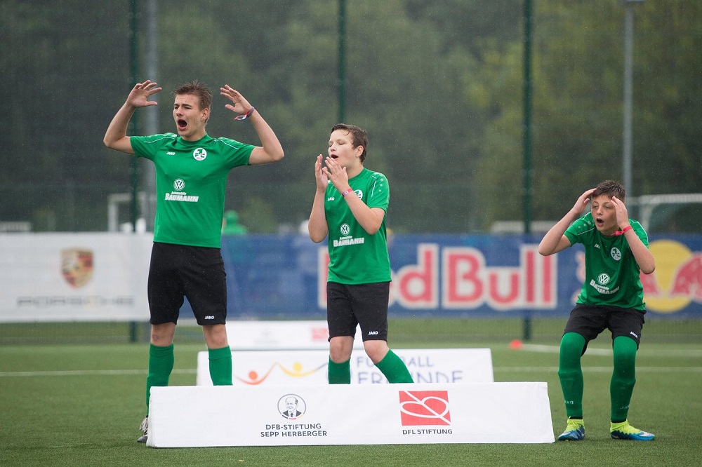 Drei begeisterte Jugendliche im grünen Fußballtrikot mit dem Volkswagen-Logo stehen auf einem Podest und jubeln in die Höhe. Im Hintergrund sind Werbebanner von Red Bull, der DFL Stiftung und der DFB-Stiftung Sepp Herberger zu sehen.