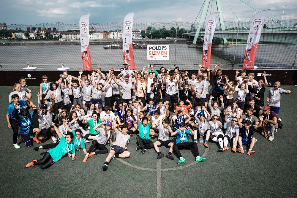Eine große Gruppe von Jugendlichen und Betreuern posiert lächelnd auf einem Kunstrasenplatz für ein Foto. Viele tragen Medaillen um den Hals. Im Hintergrund sind Banner mit dem Aufdruck "Poldi's Eleven" zu sehen, sowie eine Brücke und der Rhein.