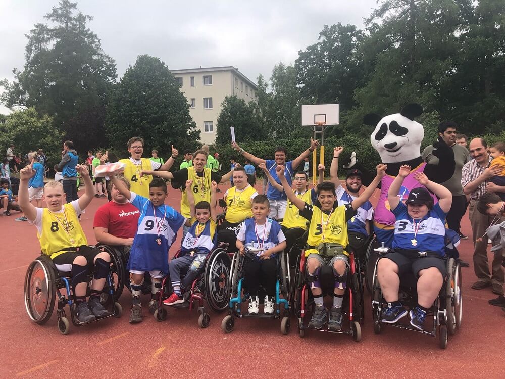 Eine Gruppe von Kindern und Jugendlichen mit und ohne Behinderung feiert auf einem Basketballplatz. Einige sitzen in Rollstühlen, andere stehen und halten die Arme in die Höhe. Im Hintergrund steht eine Person in einem Fußballmaskottchen-Kostüm.