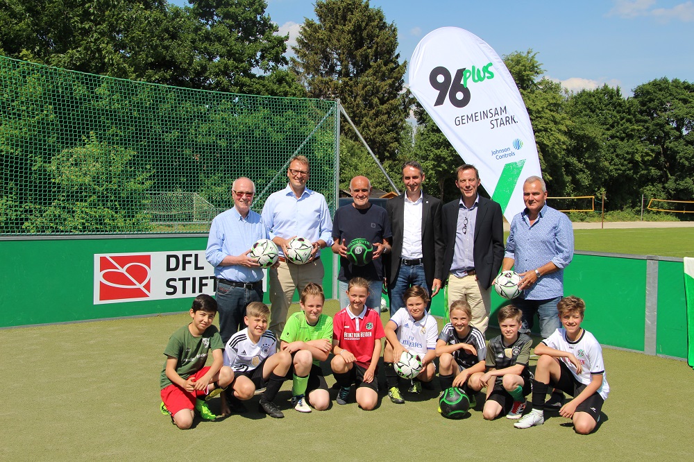 Eine Gruppe von Kindern und Männern steht vor einem grünen Kunstrasenplatz. Die Kinder, gekleidet in Sportkleidung, sitzen oder knien in der ersten Reihe. Hinter ihnen stehen Männer in Hemden und Jacken, darunter Vertreter der DFL Stiftung, 96plus und Johnson Controls. Im Hintergrund ist ein Fußballtor zu sehen.