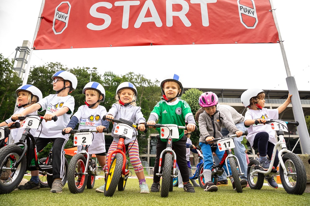 Eine Gruppe Kinder auf Laufrädern steht auf einer grünen Wiese vor einem roten Banner mit der Aufschrift "START". Die Kinder tragen Helme und Startnummern und schauen erwartungsvoll in die Kamera.