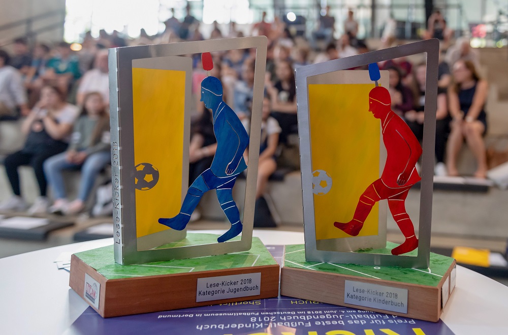 Preisverleihung "Lese-Kicker 2018" im Deutschen Fußballmuseum Dortmund: Blick auf die gläsernen Trophäen in Form von Fußballspielern, die für die Auszeichnung im Bereich Fußballjugendbuch vergeben wurden. Im Hintergrund sind angeregte Zuschauer zu sehen.