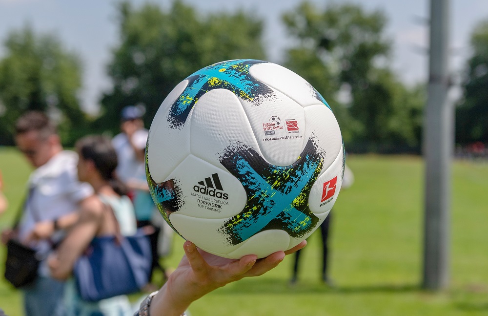 Ein Fußball mit dem Aufdruck "adidas" und dem Logo der DFL-Stiftung wird von einer Person in die Höhe geworfen. Im Hintergrund sind verschwommene Personen und eine grüne Sportfläche zu erkennen.