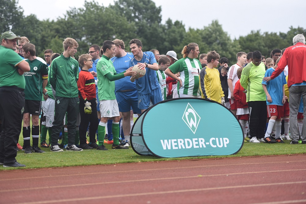 Fußballspieler in grünen und roten Trikots stehen nach einem Spiel zusammen und unterhalten sich. Im Hintergrund ein Banner mit dem Aufdruck "WERDER-CUP".