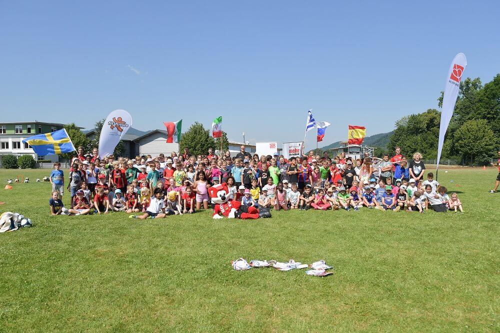 Eine große Gruppe von Kindern und Jugendlichen steht auf einem grünen Rasen und lächelt in die Kamera. Im Hintergrund sind verschiedene Flaggen und Gebäude zu sehen. Im Vordergrund steht ein Banner mit der Aufschrift "fit4future".