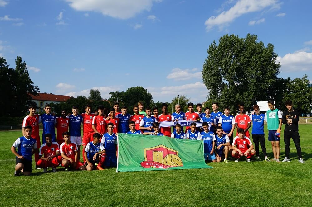 Eine Gruppe Jugendlicher im Fußballtrikot steht auf einem grünen Rasen vor einem Banner mit dem Logo von "Bielefeld United". Im Hintergrund sind Bäume und Gebäude zu sehen.