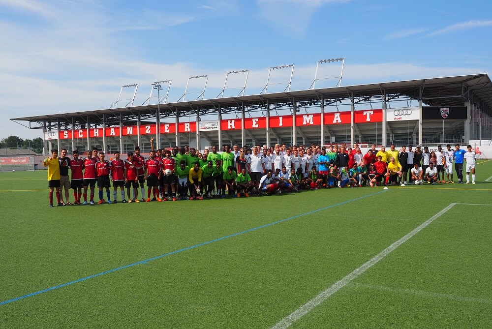 Eine große Gruppe von Kindern und Jugendlichen steht auf einem Fußballfeld vor einer Tribüne mit der Aufschrift "SCHANZER HEIMAT". Im Hintergrund ist ein Fußballstadion zu sehen.