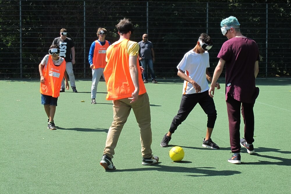 Eine Gruppe von Menschen spielt auf einem grünen Sportplatz Fußball. Einige tragen orangefarbene Westen, andere sind zivil gekleidet. Ein Spieler steht kurz davor, den Ball zu schießen. Im Hintergrund sind weitere Personen zu sehen, die am Spiel teilnehmen oder zusehen.