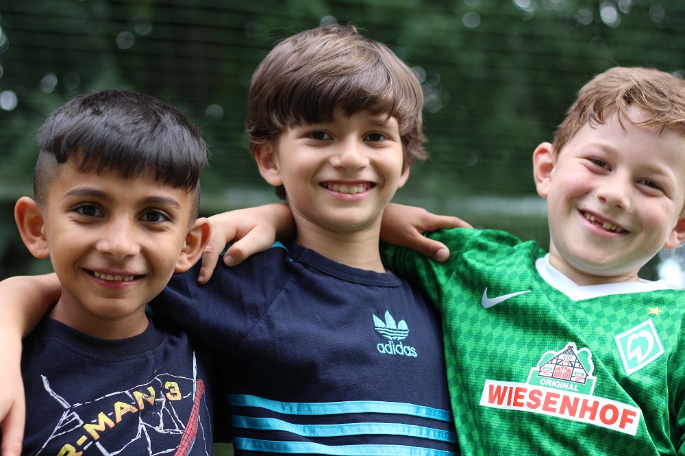 Drei lächelnde Jungen mit unterschiedlicher Herkunft umarmen sich vor grünem Hintergrund – ein Symbol für das Willkommensbündnis Bremen der DFL Stiftung.
