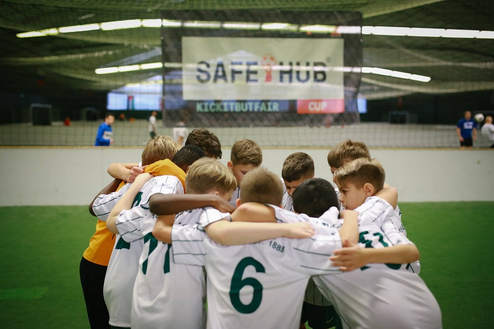 Eine Gruppe Jugendlicher in weißen Trikots mit schwarzen Details und den Rückennummern 6 und 11 steht eng zusammen im Kreis, die Arme ineinander verschlungen. Im Hintergrund ist ein Banner mit der Aufschrift "SAFE HUB" zu sehen.