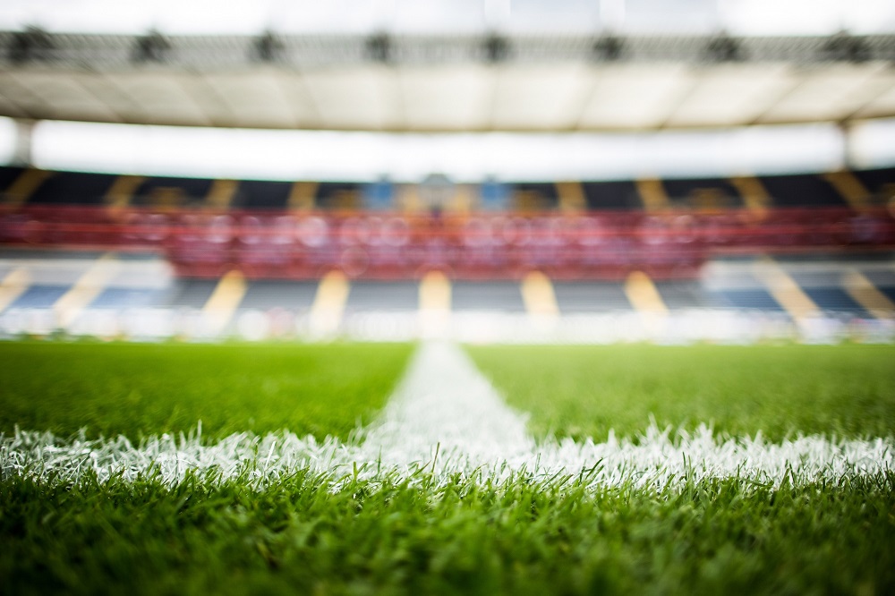 Fußballfeld mit Mittellinie, im Hintergrund verschwommen die leeren Zuschauerränge eines Stadions.