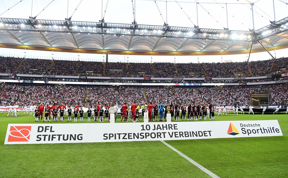 Fußballstadion mit vollem Rang, im Vordergrund Spieler in roten und weißen Trikots vor einer Werbeplane mit dem Logo der DFL Stiftung und der Deutschen Sporthilfe sowie dem Schriftzug "10 Jahre Spitzensport verbindet".